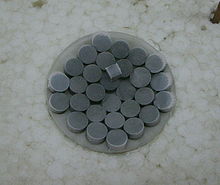 Pastillas de color gris oscuro sobre un vidrio de reloj. Una pieza cúbica del mismo material en la parte superior de las pastillas.