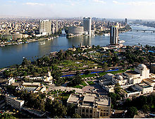 Un río azul fuerte divide el paisaje urbano, que es principalmente de vegetación y dispersos edificios de baja altura en el primer plano, y denso, con varios altos edificios modernos en el fondo