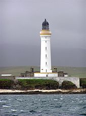 Un faro blanco con una franja marrón alrededor del parapeto y linterna de color oscuro se sienta en una costa rocosa. Una pared blanca oscurece la planta baja de edificios de piedra gris reunidos alrededor de su base.