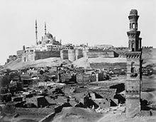Una mezquita múltiples cúpula domina la amurallada ciudadela, con tumbas en ruinas y un minarete solitario delante.