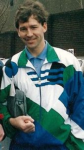Un hombre sonriente con el pelo oscuro que llevaba un chándal blanco, verde y azul arriba sobre una camisa azul. Él está sosteniendo una Washbag bajo su brazo derecho.