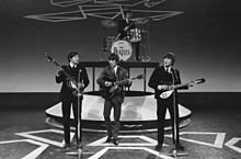 Imagen monocroma de Los Beatles tocando en un escenario con trajes oscuros.