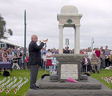 Foto de un monumento ANZAC con un hombre mayor que juega un bugle. Filas de la gente está sentada detrás del monumento. Muchas pequeñas cruces blancas con amapolas rojas se han clavado en el césped en filas a ambos lados del monumento.