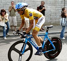 Un hombre con ropa de color amarillo y un casco azul, montando en una bicicleta. En el fondo algunos espectadores.