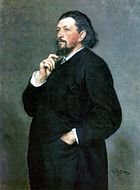 Un hombre de mediana edad con medio-largo del cabello oscuro y una barba, vestido con un traje oscuro, con una mano en el bolsillo del pantalón y la otra mano en el mentón