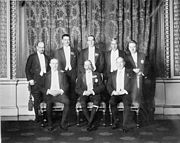 Un grupo pose de ocho hombres en trajes de noche inteligente. El rey se sienta en el centro rodeado por sus principales ministros.