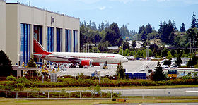 Fábrica de aviones. Edificio rectangular con seis puertas, una de ellas se abren para revelar un avión emergente. El fondo es un bosque; vehículos pequeños y calles de rodaje rodean la fábrica.