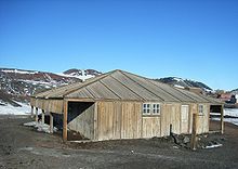 Estructura de madera con puerta y dos ventanas pequeñas. A la izquierda hay un cobertizo abierto. En el fondo son las montañas cubiertas de nieve en parte.