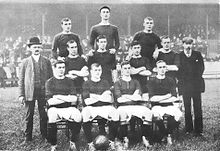 Una fotografía en blanco y negro de un equipo de fútbol en fila antes de un partido. Cuatro jugadores, vestidos con camisas oscuras, pantalones cortos ligeros y calcetines oscuros, están sentados. Cuatro jugadores más están de pie inmediatamente detrás de ellos, y tres más están de pie en un nivel más alto en la última fila. Dos hombres de traje están de pie a cada lado de los jugadores.