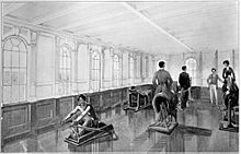 Vista del interior de un gimnasio bien iluminado. Un hombre está utilizando una máquina de remo en el primer plano, mientras que un hombre y una mujer ride máquinas de ejercicio de caballos. Otros dos hombres están de pie en el fondo visible.