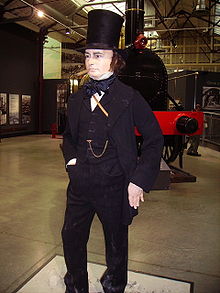 Dentro de un gran edificio, una figura de tamaño natural de un hombre de unos cincuenta años, vestido con chaqueta siglo XIX, pantalones y chaleco, con una destacada cadena de reloj sobre el pecho. Él lleva un sombrero negro alto tubo de chimenea, tiene patillas largas y tiene un cigarro en la boca. Una vieja locomotora de vapor en el fondo.