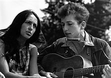 Dylan está sentado, cantando y tocando la guitarra. Sentado a su derecha es una mujer mirando hacia arriba y cantar con él.